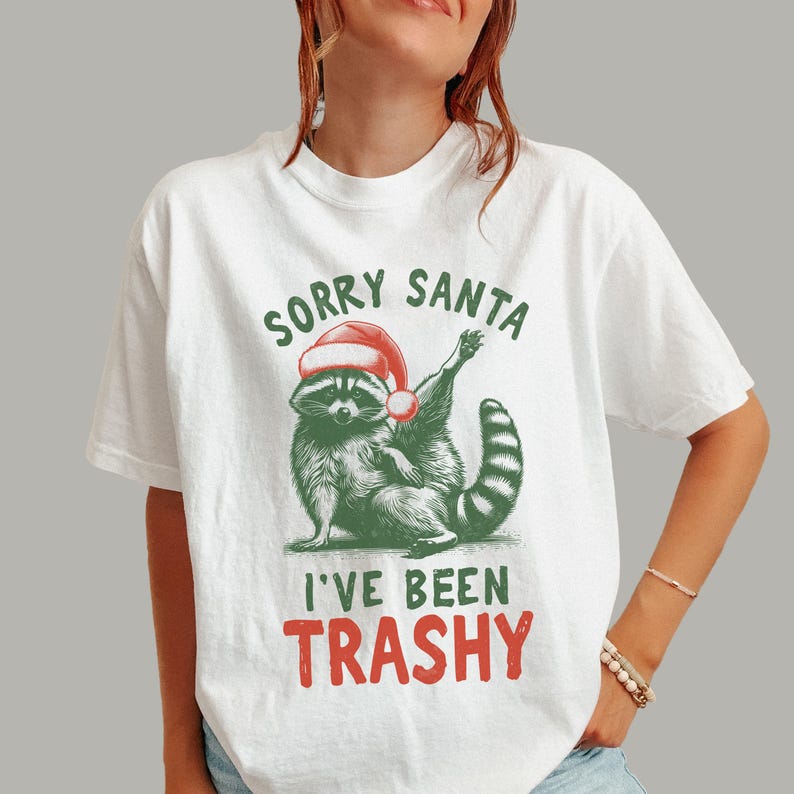 May include: White t-shirt featuring a raccoon wearing a Santa hat. The raccoon is illustrated in green and black, with the text "SORRY SANTA I'VE BEEN TRASHY" in green and red. The shirt is a casual, festive design.
