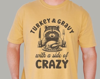 May include: Mustard yellow t-shirt with a graphic featuring a raccoon holding a knife and fork, with a roasted turkey on a plate. The text reads "TURKEY & GRAVY" and "with a side of CRAZY".