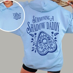 Puede incluir: Sudadera con capucha azul claro con cord&oacute;n. El lado izquierdo del pecho presenta un peque&ntilde;o gr&aacute;fico azul oscuro de una planchette. La parte posterior de la sudadera tiene el texto "Summoning a Shadow Daddy" sobre un gr&aacute;fico de planchette con las palabras "Yes Sir" y "Good Girl".