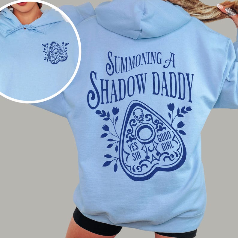 Puede incluir: Sudadera con capucha azul claro con cord&oacute;n. El lado izquierdo del pecho presenta un peque&ntilde;o gr&aacute;fico azul oscuro de una planchette. La parte posterior de la sudadera tiene el texto "Summoning a Shadow Daddy" sobre un gr&aacute;fico de planchette con las palabras "Yes Sir" y "Good Girl".