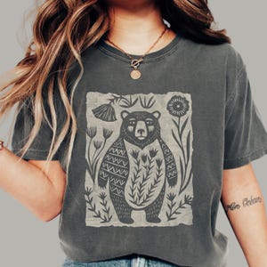 May include: A charcoal gray short-sleeve t-shirt featuring a central graphic print of a bear surrounded by stylized flowers and foliage. The bear has a patterned chest and arms, and the design is set against a light gray rectangular background.