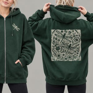 May include: A dark forest green zip-up hoodie featuring a small dragonfly graphic on the left chest. The back displays a large, square, light-colored patch with a detailed black ink-style illustration of two dragonflies surrounded by various flowers.
