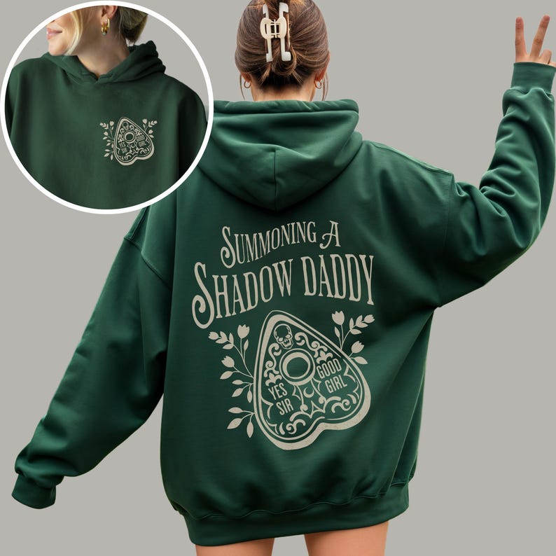 Puede incluir: Sudadera con capucha verde oscuro con el texto "Summoning A Shadow Daddy" en una fuente estilizada en la espalda. Un dise&ntilde;o de planchette est&aacute; debajo del texto. La parte delantera de la sudadera tiene un dise&ntilde;o de planchette m&aacute;s peque&ntilde;o.