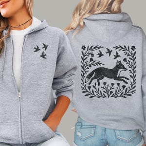 May include: A heather gray zip-up hoodie featuring a black graphic of three small birds on the front left chest. The back displays a large black graphic of a fox running within a square frame of stylized leaves, flowers, and three birds.