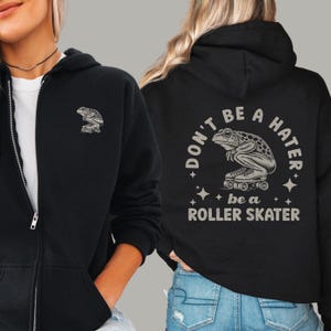 May include: A black zip-up hoodie featuring a small graphic of a frog on roller skates on the front left chest. The back displays a larger version of the same frog graphic with the text "DON'T BE A HATER be a ROLLER SKATER" in light gray.