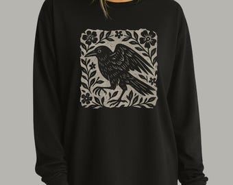 May include: A black long-sleeve sweatshirt featuring a square graphic print on the chest. The print displays a black crow standing among stylized leaves and flowers against a light gray background, rendered in a block-print style.
