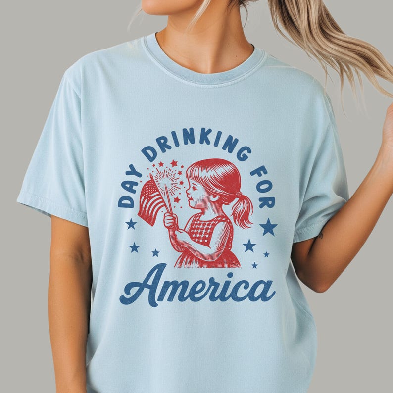 May include: Light blue t-shirt with a graphic featuring a young girl holding a sparkler and an American flag. The text reads "Day Drinking for America" in a circular design with stars.