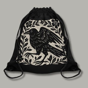 May include: Black drawstring backpack featuring a detailed black and white illustration of a raven surrounded by floral and leaf elements. The raven is depicted with its wing outstretched, creating a striking design.