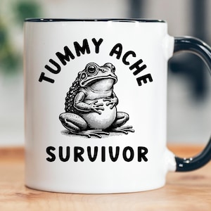 May include: White ceramic mug with a black rim and handle. The mug features a black and white illustration of a frog holding its belly, with the words "TUMMY ACHE" and "SURVIVOR" in black lettering.