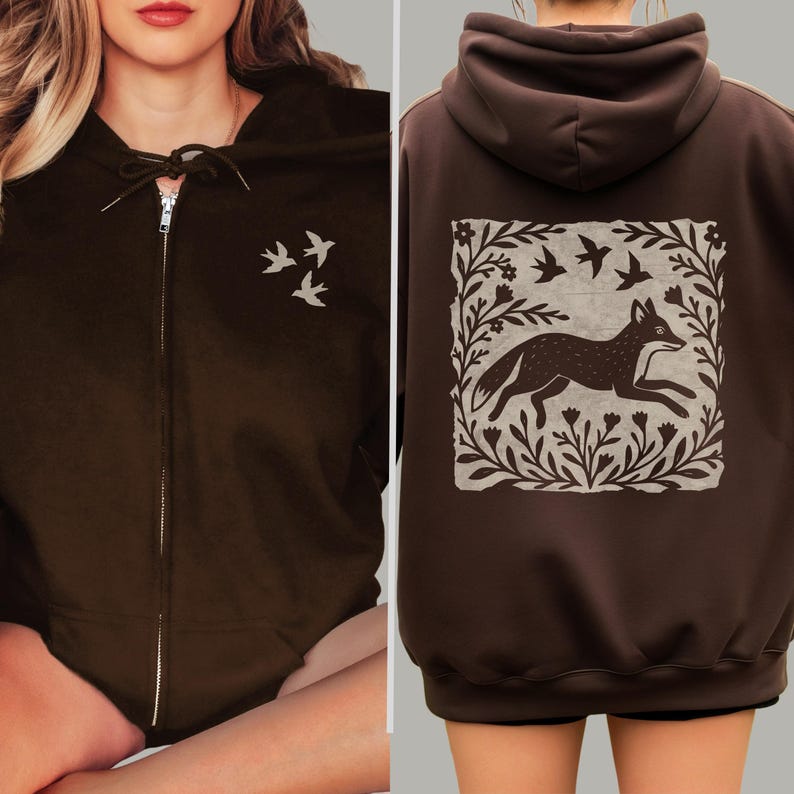 May include: A dark brown zip-up hoodie featuring a small graphic of three birds on the front left chest. The back of the hoodie displays a large, square-shaped print of a fox running through a field of flowers and foliage, surrounded by two birds.