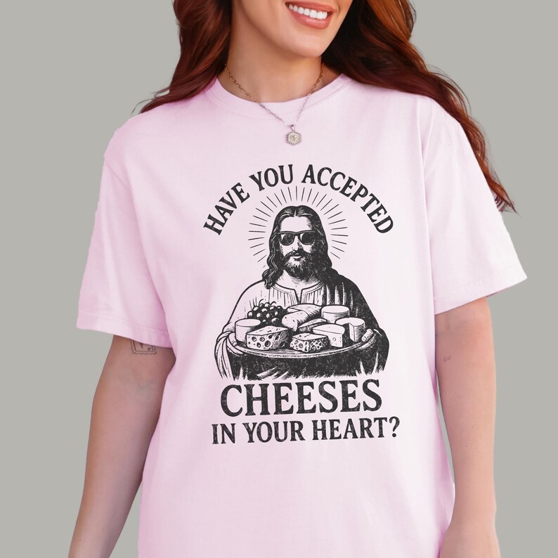 Pu&ograve; includere: Una t-shirt a maniche corte rosa chiaro con una grafica nera raffigurante una figura barbuta con occhiali da sole che tiene in mano un vassoio di formaggi vari. Il testo sopra e sotto la grafica recita: "HAVE YOU ACCEPTED CHEESES IN YOUR HEART?"