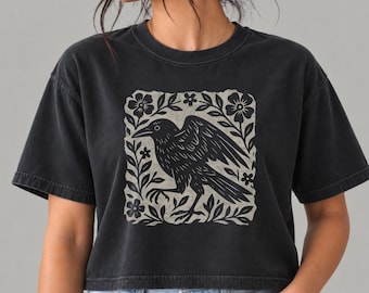 May include: A charcoal black short-sleeve t-shirt featuring a centered square graphic print. The design displays a black crow in profile with wings partially spread, surrounded by stylized floral and leaf motifs in a light beige color.
