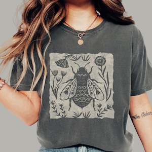 May include: A charcoal gray short-sleeve t-shirt featuring a central square graphic of a stylized bee surrounded by various botanical elements. The bee design includes intricate geometric patterns on its wings and body, rendered in a dark ink color.