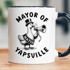 May include: White ceramic mug with a black rim and handle. The mug features a cartoon illustration of a goose wearing a top hat and a "MAYOR" sash, holding a megaphone. Text on the mug reads "MAYOR OF YAPSVILLE."