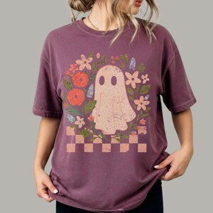 May include: A maroon t-shirt featuring a ghost design surrounded by flowers. The ghost is light peach-colored with two oval eyes. The flowers are orange, pink, and blue. A checkered pattern is at the bottom of the design.