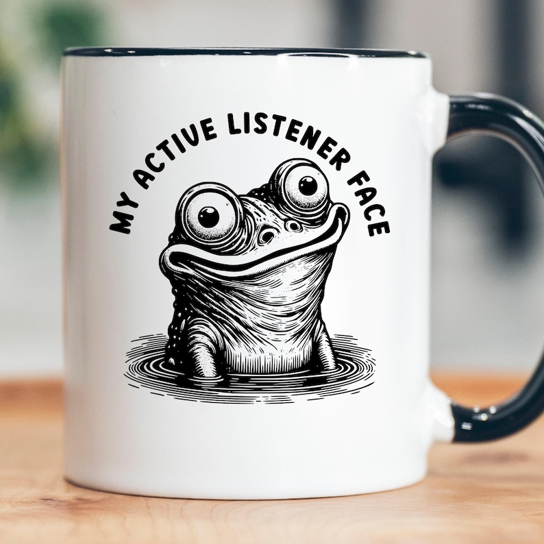 Puede incluir: Taza de cer&aacute;mica blanca con asa y borde negros. La taza presenta una ilustraci&oacute;n en blanco y negro de una rana con ojos grandes y el texto "MY ACTIVE LISTENER FACE" en un dise&ntilde;o circular alrededor de la rana.