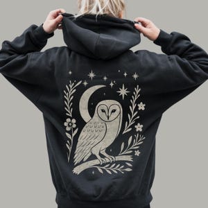 May include: A black hooded sweatshirt featuring a large, light-colored graphic on the back. The design depicts a barn owl perched on a branch, framed by a crescent moon, small flowers, leafy sprigs, and various four-pointed stars.