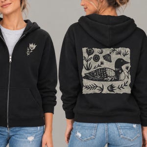 May include: A black zip-up hoodie featuring a small light-colored flower graphic on the left chest. The back displays a large, square, light-colored block print design of a loon surrounded by stylized floral and leaf motifs in black ink.