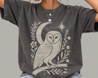 May include: A charcoal gray short-sleeve t-shirt featuring a light-colored graphic print of an owl perched on a branch. The design includes a crescent moon, a star, and floral elements surrounding the owl. The shirt has a crew neckline.