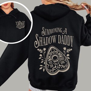 Puede incluir: Sudadera con capucha negra con un gr&aacute;fico de tabla Ouija beige en la parte delantera y trasera. La parte trasera de la sudadera con capucha presenta el texto "Summoning a Shadow Daddy" encima del dise&ntilde;o de la tabla Ouija. El dise&ntilde;o incluye las palabras "Yes Sir" y "Good Girl".