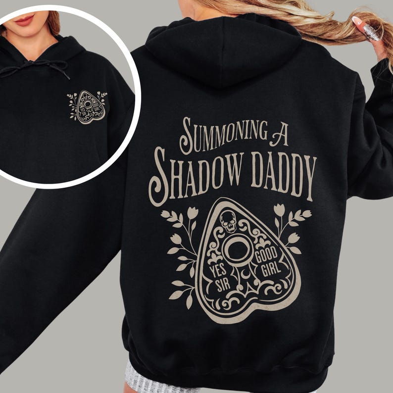 Puede incluir: Sudadera con capucha negra con un gr&aacute;fico de tabla Ouija beige en la parte delantera y trasera. La parte trasera de la sudadera con capucha presenta el texto "Summoning a Shadow Daddy" encima del dise&ntilde;o de la tabla Ouija. El dise&ntilde;o incluye las palabras "Yes Sir" y "Good Girl".
