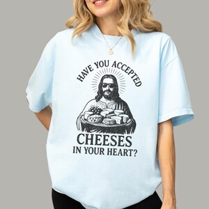 Pu&ograve; includere: Una t-shirt azzurra con una stampa grafica nera di una figura barbuta con occhiali da sole che tiene in mano un vassoio di vari formaggi. Il testo sopra e sotto la grafica recita: "HAVE YOU ACCEPTED CHEESES IN YOUR HEART?"