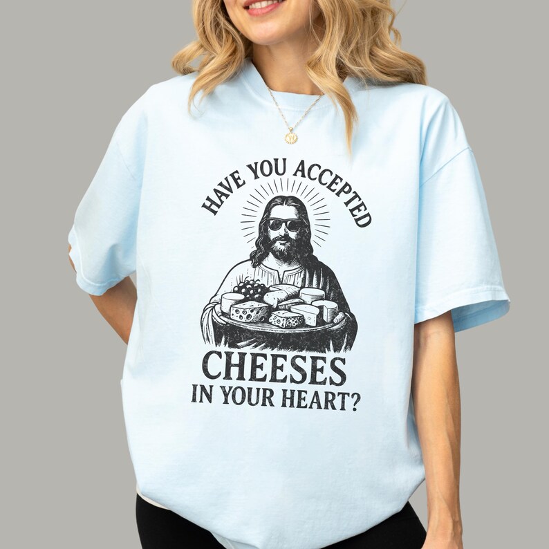 Pu&ograve; includere: Una t-shirt azzurra con una stampa grafica nera di una figura barbuta con occhiali da sole che tiene in mano un vassoio di vari formaggi. Il testo sopra e sotto la grafica recita: "HAVE YOU ACCEPTED CHEESES IN YOUR HEART?"
