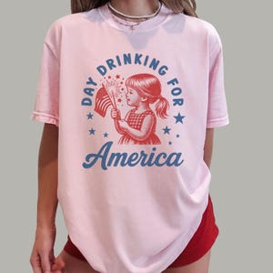 May include: A light pink t-shirt with a graphic design. The design features a vintage illustration of a girl holding a sparkler and an American flag, with the words "Day Drinking for America" in blue.
