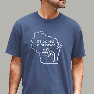 May include: A navy blue short-sleeved t-shirt featuring a white outline of the state of Wisconsin. Inside the outline, the text reads 'It's called a bubbler' above a white line-art icon of a person drinking from a water fountain.