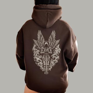 Op de afbeelding: Een bruine hoodie met een gedetailleerde afbeelding van een engel met grote vleugels, een zwaard en een halo. Het engelontwerp is in grijstinten en wit, tegen een bruine achtergrond. De hoodie heeft een relaxte pasvorm.
