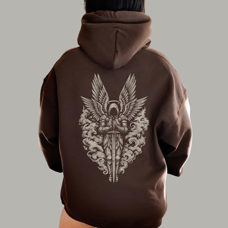Op de afbeelding: Een bruine hoodie met een gedetailleerde afbeelding van een engel met grote vleugels, een zwaard en een halo. Het engelontwerp is in grijstinten en wit, tegen een bruine achtergrond. De hoodie heeft een relaxte pasvorm.