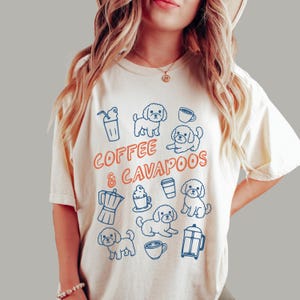 May include: Cream-colored t-shirt with the words "Coffee & Cavapoos" in orange and blue. The shirt features blue line drawings of coffee-related items and Cavapoo dogs. The design includes coffee cups, a French press, and a to-go cup.