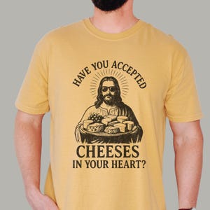 Pu&ograve; includere: Una t-shirt giallo senape con una stampa grafica nera di una figura barbuta con occhiali da sole, che regge un vassoio di vari formaggi. Il testo sopra recita "HAVE YOU ACCEPTED" e il testo sotto recita "CHEESES IN YOUR HEART?"