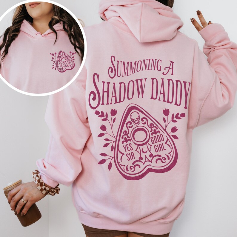 Puede incluir: Sudadera con capucha rosa claro con un dise&ntilde;o de tablero Ouija rosa oscuro en la parte delantera y trasera. La parte trasera presenta el texto "Summoning a Shadow Daddy" encima del tablero, con "Yes Sir" y "Good Girl" en el tablero.