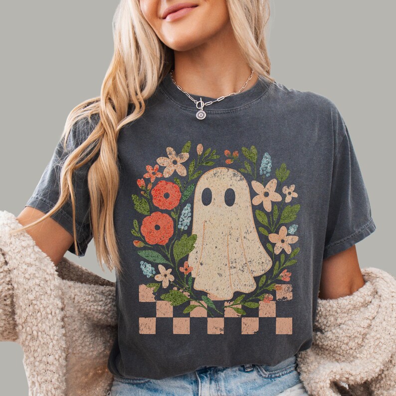 May include: Dark gray t-shirt featuring a ghost illustration surrounded by a floral wreath with orange, pink, and blue flowers. A checkered pattern is at the bottom of the design. The shirt has a vintage, worn look.