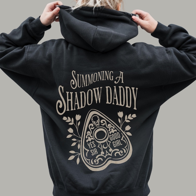 Puede incluir: Sudadera con capucha negra con el texto "Summoning A Shadow Daddy" en una fuente estilizada. Debajo del texto hay un dise&ntilde;o de plancheta beige con las palabras "Yes Sir" y "Good Girl". La sudadera tiene una capucha con cord&oacute;n.