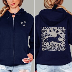 May include: A navy blue zip-up hoodie featuring a small graphic of three birds on the front left chest. The back displays a large square print of a fox surrounded by a floral border and birds, rendered in a light gray color on the dark fabric.