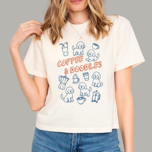 May include: A cream-colored short-sleeve t-shirt featuring a graphic print in blue and orange. The design includes the text COFFEE & DOODLES surrounded by illustrations of various coffee drinks and several small doodle dog figures.