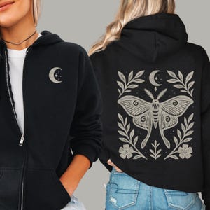 May include: Black zip-up hoodie with a small crescent moon design on the chest. The back of the hoodie features a large moth illustration with a crescent moon, floral, and leaf design in a neutral color.