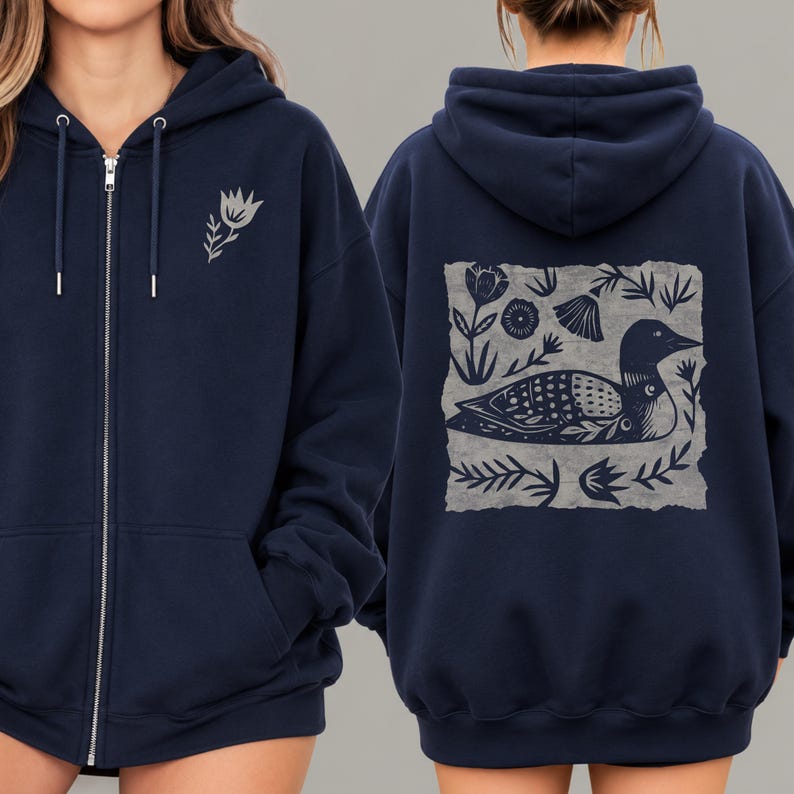 May include: A navy blue zip-up hoodie featuring a small light gray flower graphic on the front left chest. The back displays a large square light gray print of a loon bird surrounded by various botanical elements and stylized floral patterns.