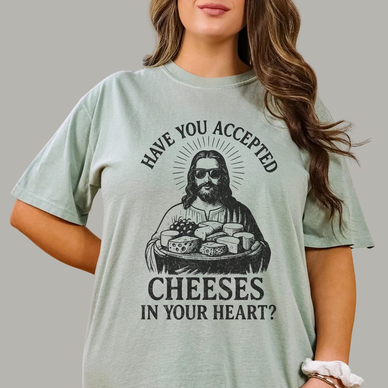 Pu&ograve; includere: Una t-shirt a maniche corte color verde salvia chiaro con una stampa grafica nera di una figura barbuta con occhiali da sole che tiene in mano un vassoio di formaggi. Il testo sopra e sotto la grafica recita, Have you accepted cheeses in your heart?