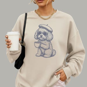 May include: A beige crewneck sweatshirt featuring a blue graphic illustration of a poodle wearing a beret and round sunglasses while holding a coffee cup. The sweatshirt has long sleeves and a ribbed neckline, worn with a black shoulder bag.