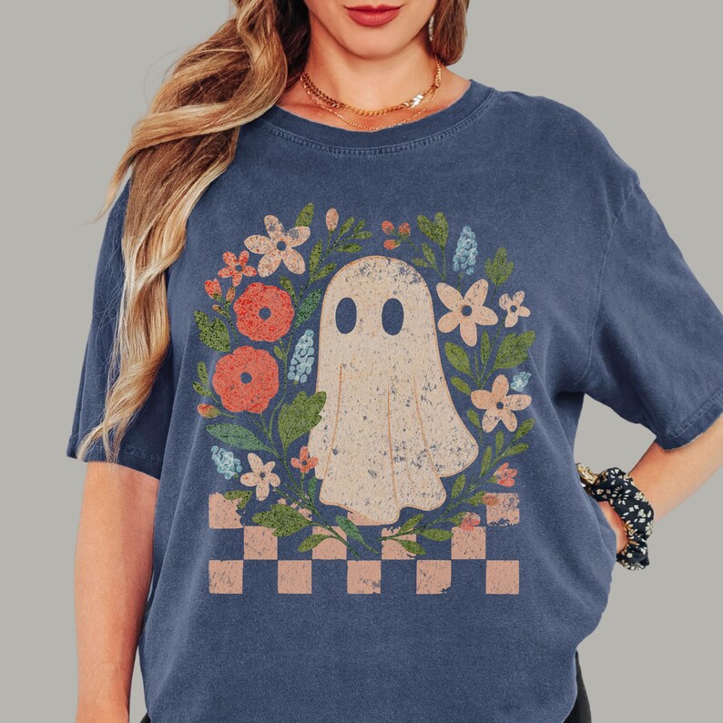 May include: A navy blue t-shirt featuring a floral wreath design with a ghost in the center. The wreath includes orange, pink, and blue flowers, with a checkered pattern at the bottom. The shirt has a vintage, distressed look.