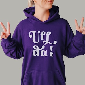 May include: A deep purple long-sleeve hooded sweatshirt featuring a front kangaroo pocket. The center of the chest displays the white text Uff da! with the exclamation point stylized as a small outline of the state of Minnesota.