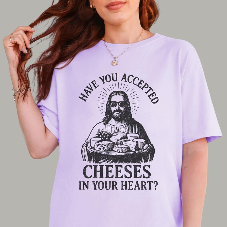 Pu&ograve; includere: Una t-shirt a maniche corte color viola chiaro con una stampa grafica nera di una figura barbuta con occhiali da sole che tiene in mano un vassoio di formaggi. Il testo recita: Have you accepted cheeses in your heart? in carattere nero.