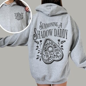 Puede incluir: Una sudadera con capucha gris claro con un dise&ntilde;o de tablero Ouija negro en la parte delantera y trasera. La parte trasera presenta el texto "Summoning a Shadow Daddy" encima del tablero. El tablero incluye las frases "Yes Sir" y "Good Girl".