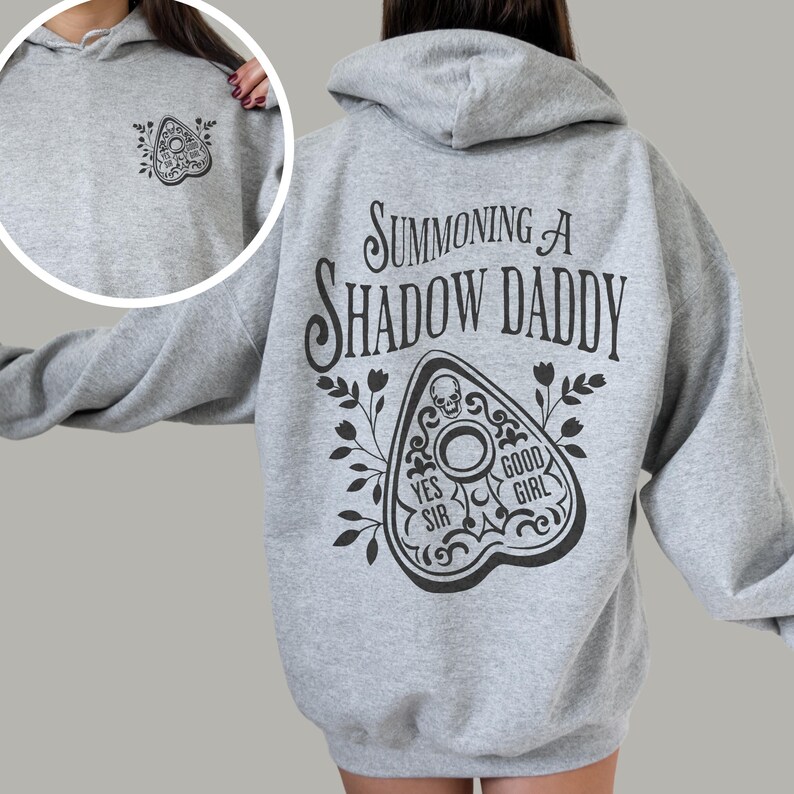 Puede incluir: Una sudadera con capucha gris claro con un dise&ntilde;o de tablero Ouija negro en la parte delantera y trasera. La parte trasera presenta el texto "Summoning a Shadow Daddy" encima del tablero. El tablero incluye las frases "Yes Sir" y "Good Girl".