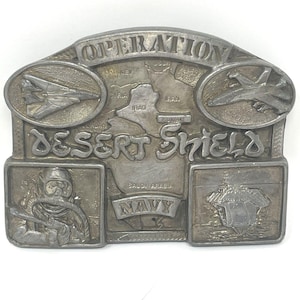 May include: Silver-toned metal belt buckle with the words "OPERATION DESERT SHIELD" and "NAVY." The buckle features images of aircraft, a map, a diver, and a ship. The buckle is rectangular with an arched top.