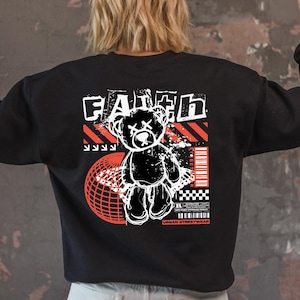 May include: Black sweatshirt with a graphic design on the back featuring a teddy bear, the word "FAITH", and a red and white globe. The design has a streetwear aesthetic.