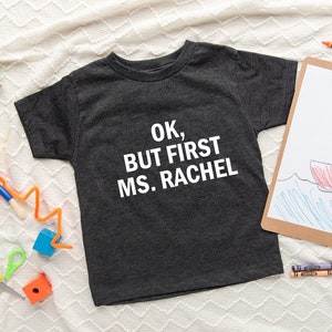 May include: A dark gray t-shirt with the text "OK, BUT FIRST MS. RACHEL" printed in white. The shirt is surrounded by colorful art supplies, including crayons, paintbrushes, and wooden blocks.
